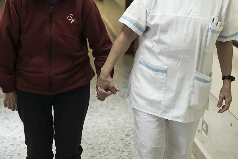 Un'anziana e un'infermiera camminano in una struttura della Sarah cooperativa, tenendosi per mano